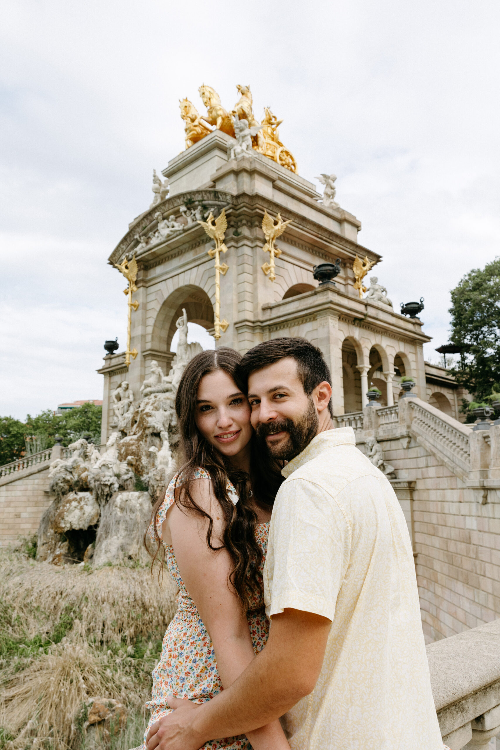 A couple embraces and smiles at the camera in front of an ornate stone fountain and arch topped with golden statues.