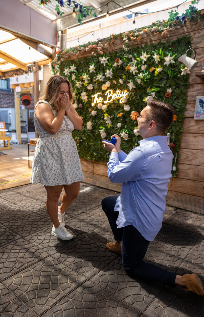 A man kneels on one knee holding an open ring box, proposing to a surprised woman who covers her mouth with both hands. They stand in a bright, decorated indoor-outdoor space with a green floral wall behind them and a glowing “To Betsy” sign, suggesting a romantic, personalized proposal setting.