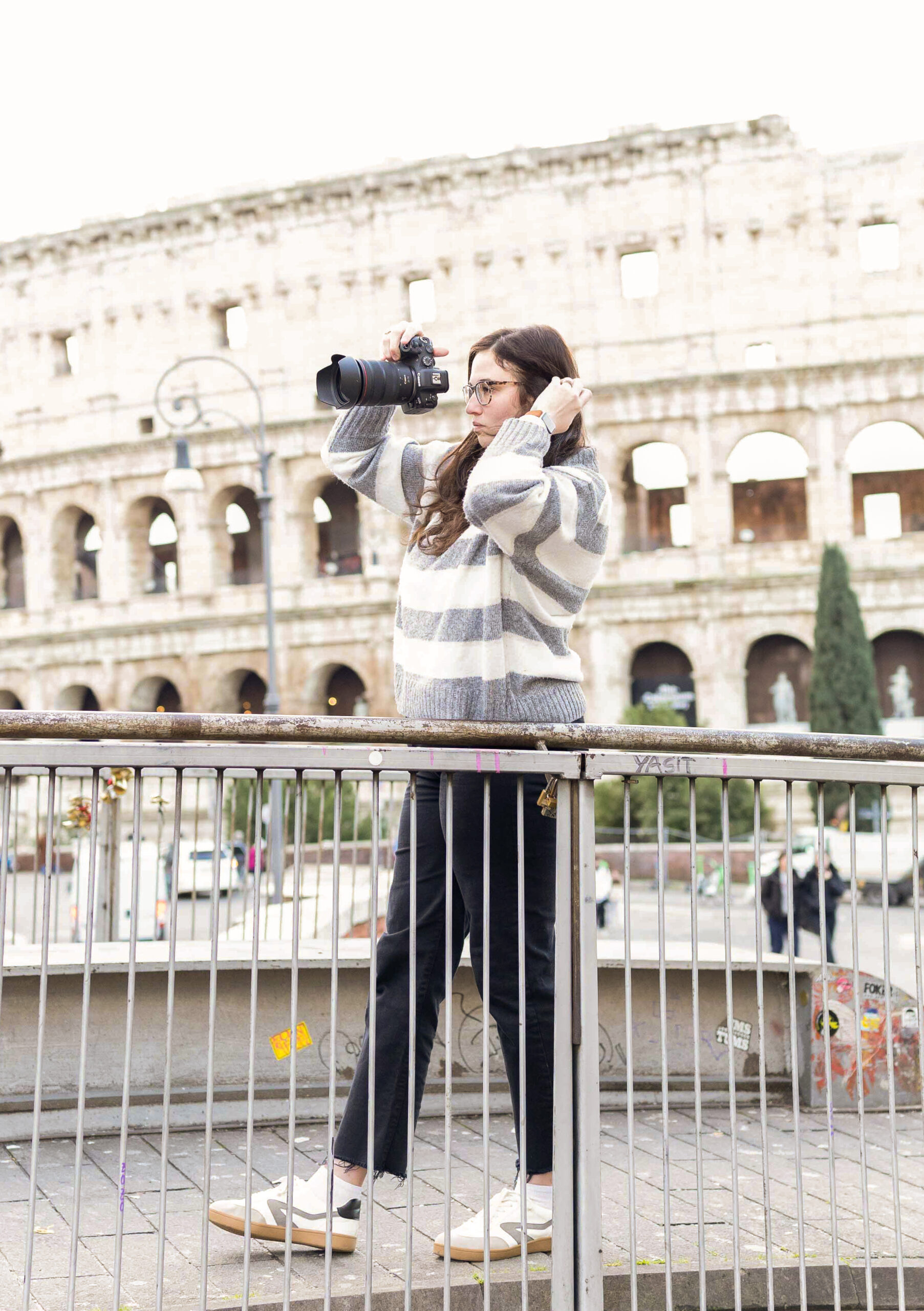A woman wearing glasses holds up a camera to take a photo while standing by a railing with the Colosseum in the background.