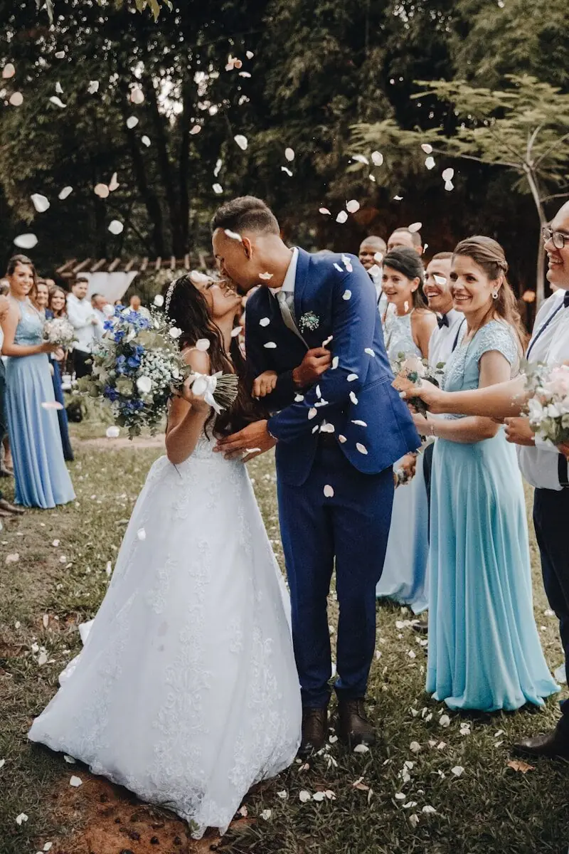 A joyful bride and groom share a kiss while walking through a shower of white flower petals, surrounded by smiling wedding guests in an outdoor garden setting in Washington DC
