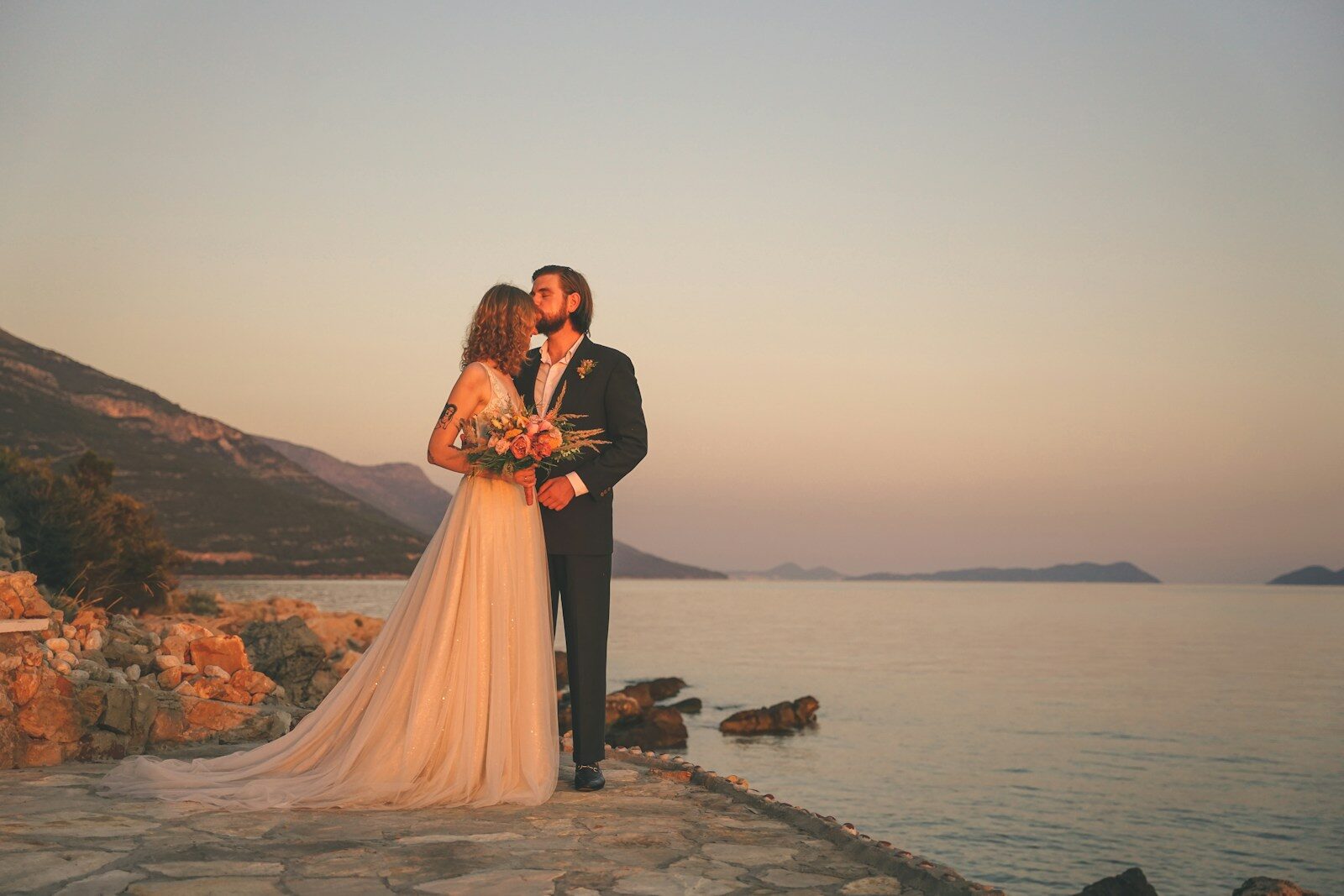A groom kisses his bride’s forehead on a stone terrace at sunset, with a calm sea and mountainous coastline stretching into the golden horizon.