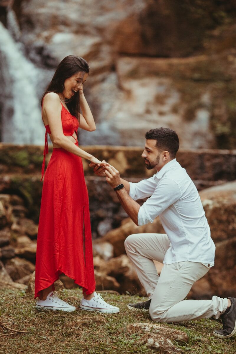 A man kneels to propose to a woman in a red dress as they smile at each other near a waterfall.