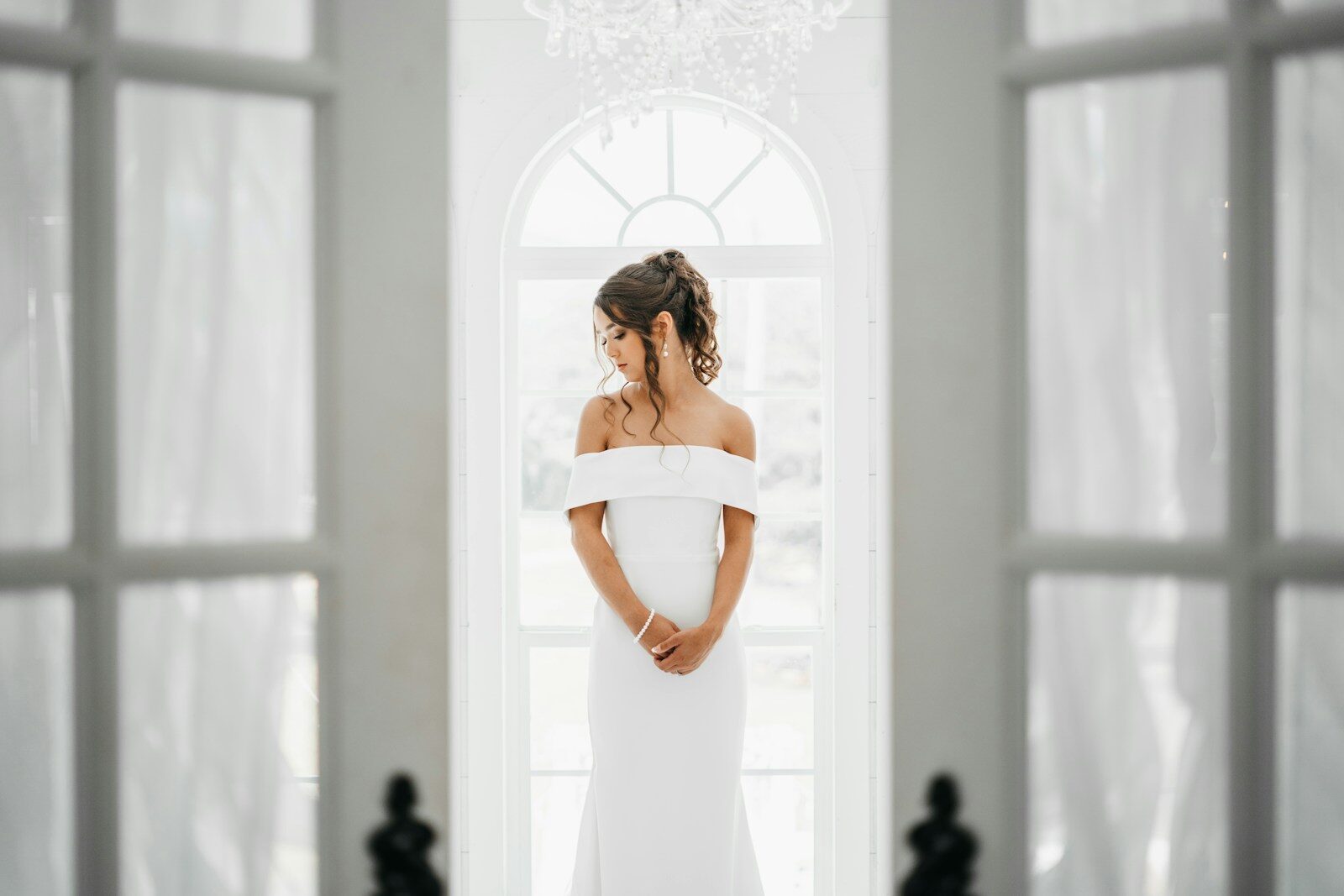 Framed by open glass doors, a bride in a minimalist off-the-shoulder gown stands pensively before a large arched window under a crystal chandelier.